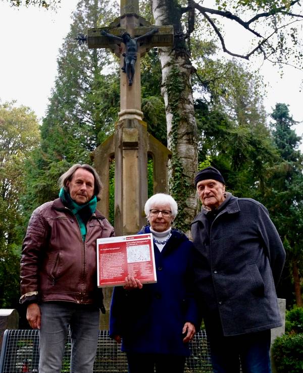 Zwei Männer und eine Frau zeigen eine rote Infotafel, die den Müngersdorfer Friedhof als Denkmal ausweist.