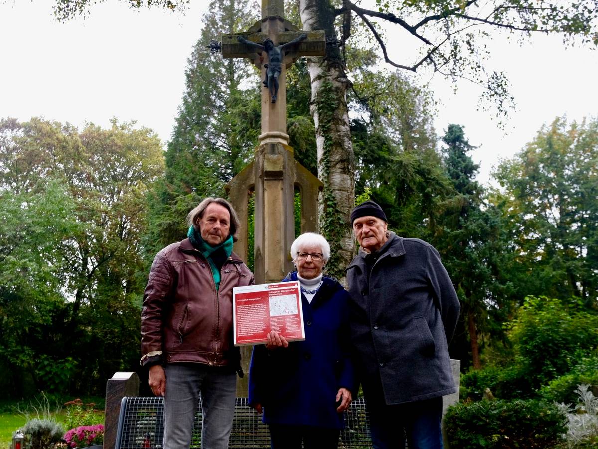 Zwei Männer und eine Frau zeigen eine rote Infotafel, die den Müngersdorfer Friedhof als Denkmal ausweist.