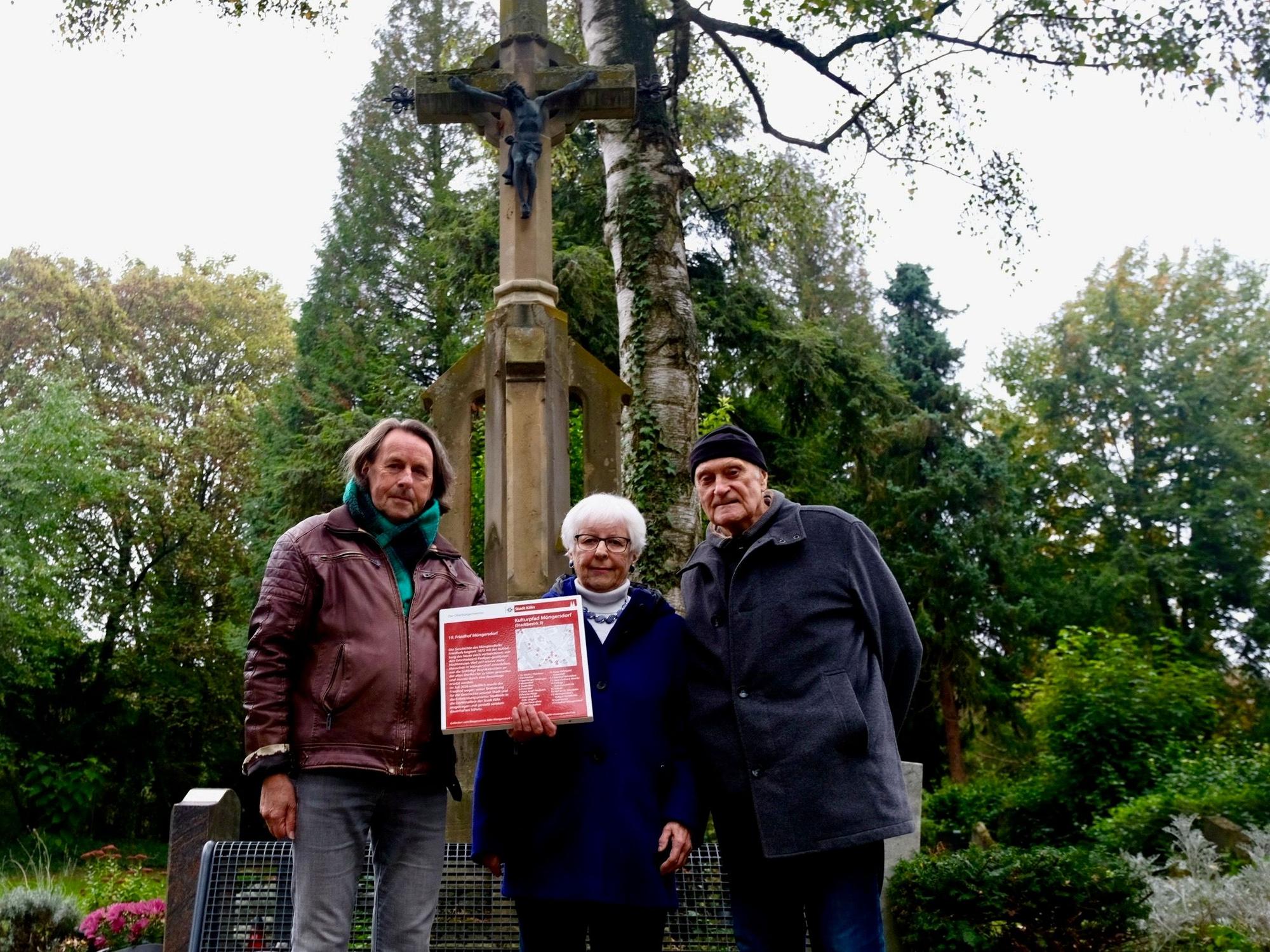 Zwei Männer und eine Frau zeigen eine rote Infotafel, die den Müngersdorfer Friedhof als Denkmal ausweist.