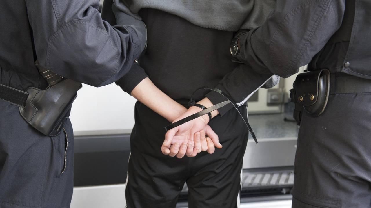 Zwei Einsatzkräfte führen einen Mann ab, dessen Hände auf dem Rücken gefesselt sind.