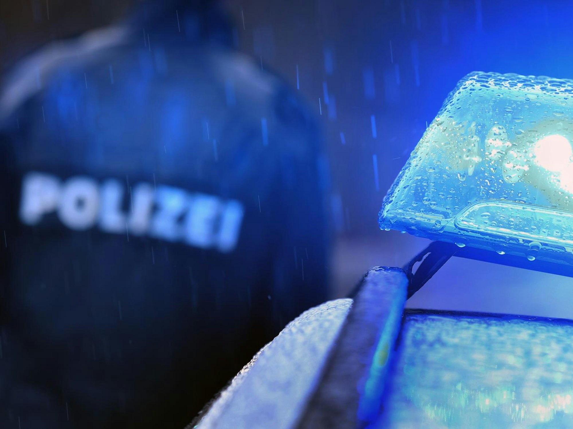 Blaulicht an einem Auto der Polizei. Im Hintergrund: eine Person, die eine Polizei-Jacke trägt.