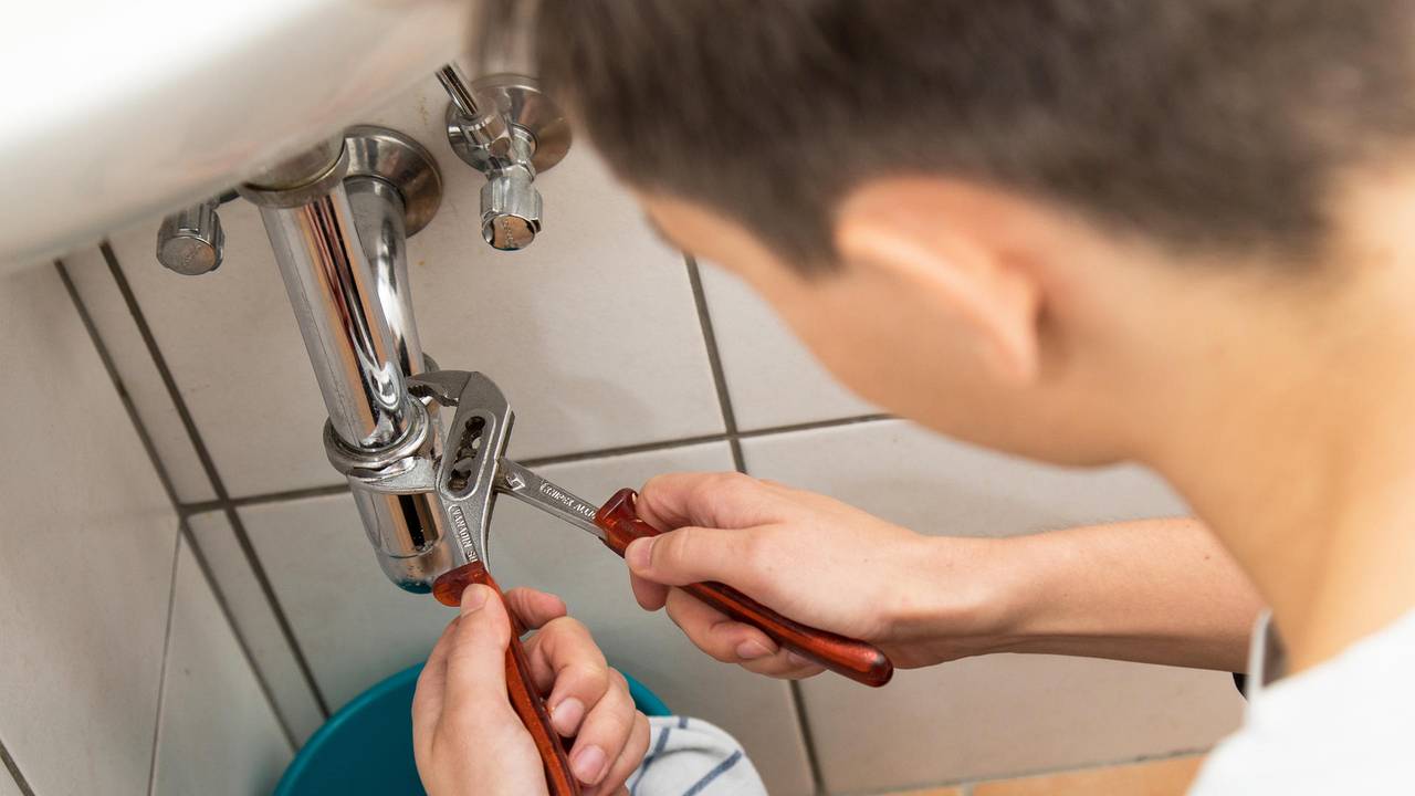 Das Symbolfoto zeigt einen Handwerker an einem Waschbeckenrohr.