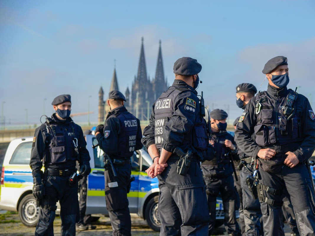 Mehrere Polizisten stehen vor mehreren Streifenwagen, im Hintergrund der Kölner Dom.