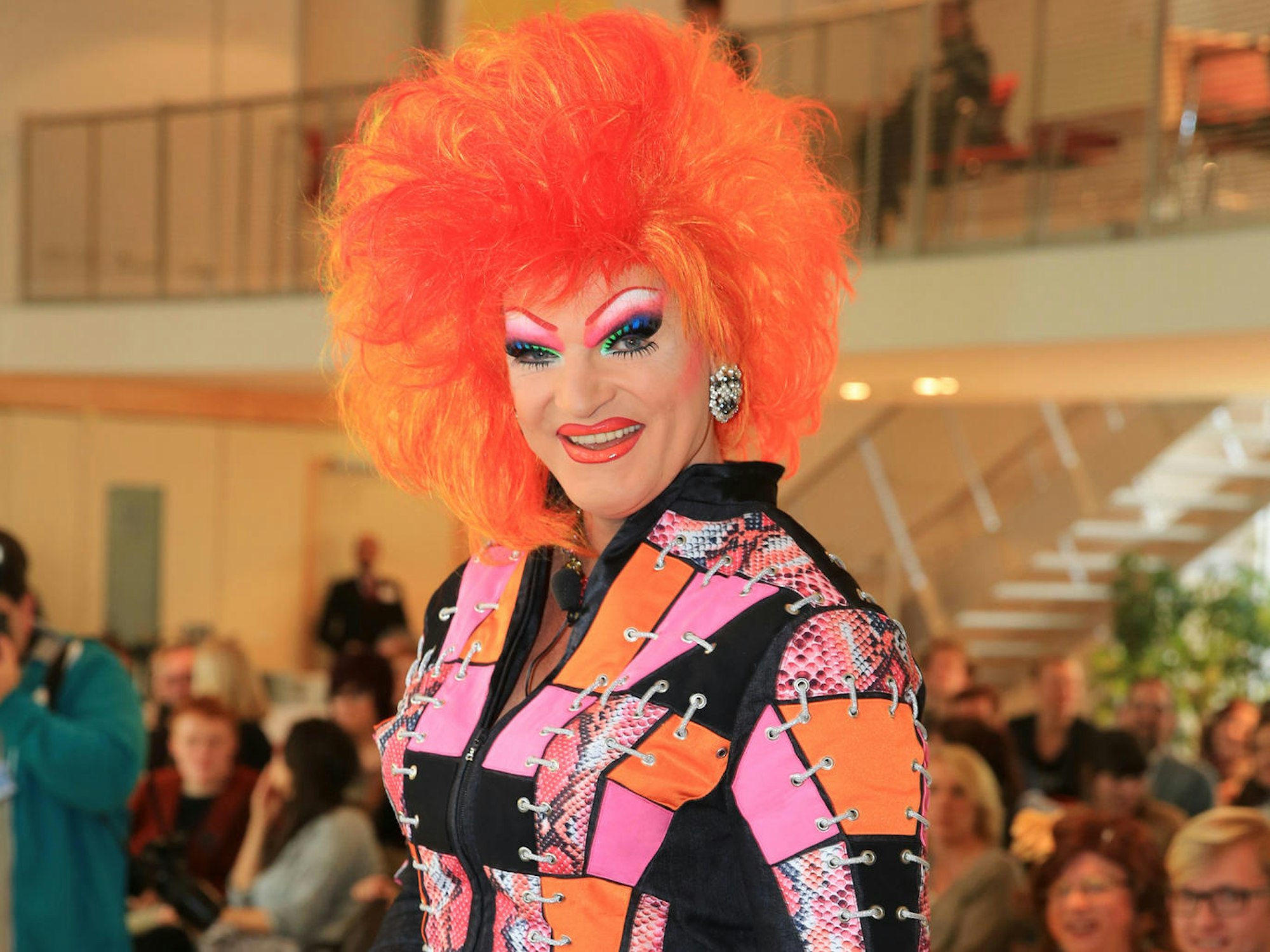 Eine stark geschminkte Person trägt eine orangefarbene Perücke und steht vor sitzendem Publikum.