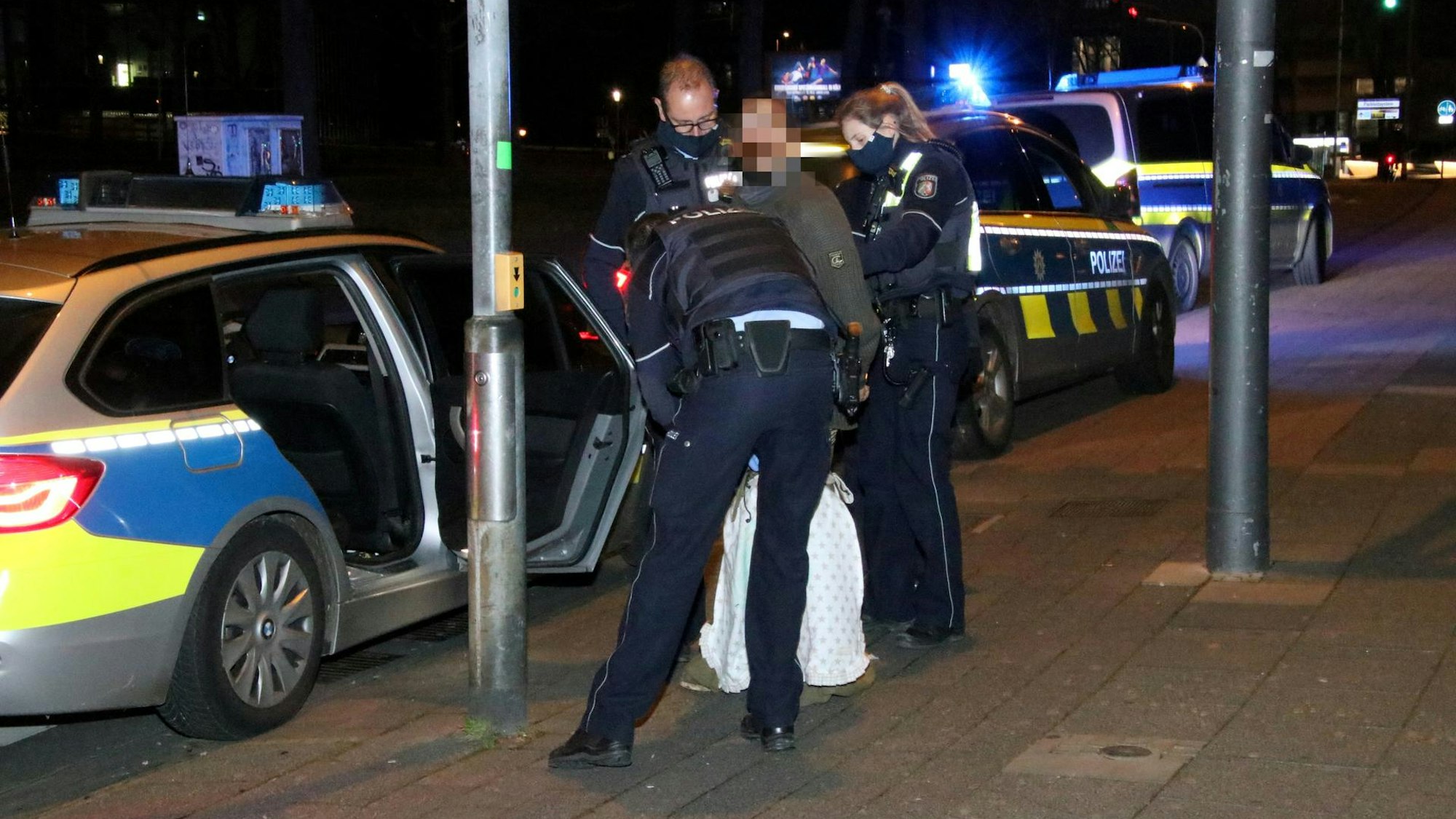 Eine Polizistin und zwei Polizisten haben einen Mann festgenommen. Sie stehen auf dem Gehweg, an dem ein Streifenwagen parkt.