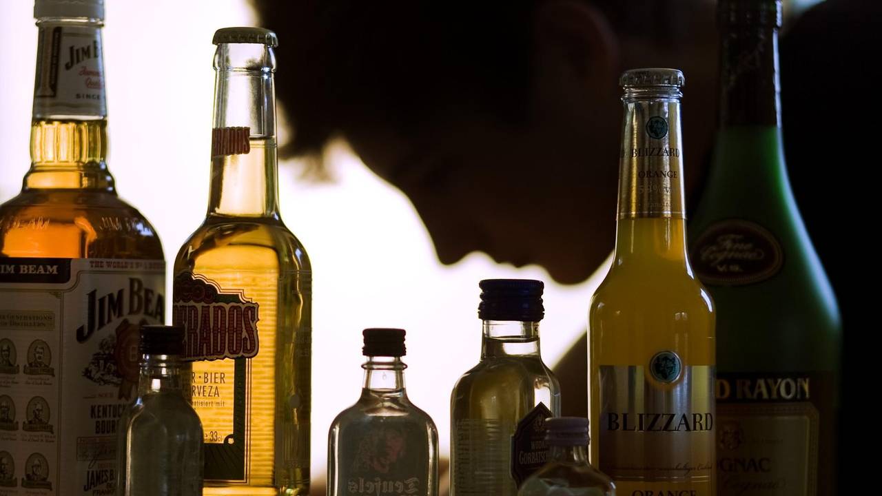 Flaschen mit Alkohol, darunter Bier und Whiskey, stehen aufgereiht vor einer Person, die nur im Profil zu sehen ist.