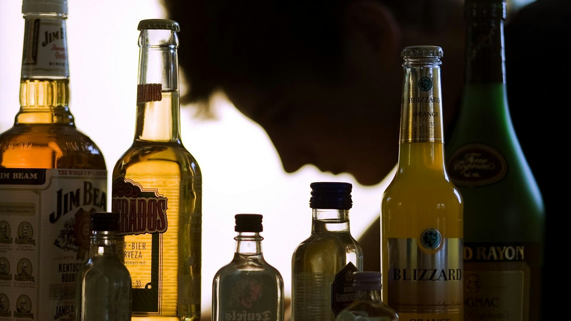 Flaschen mit Alkohol, darunter Bier und Whiskey, stehen aufgereiht vor einer Person, die nur im Profil zu sehen ist.
