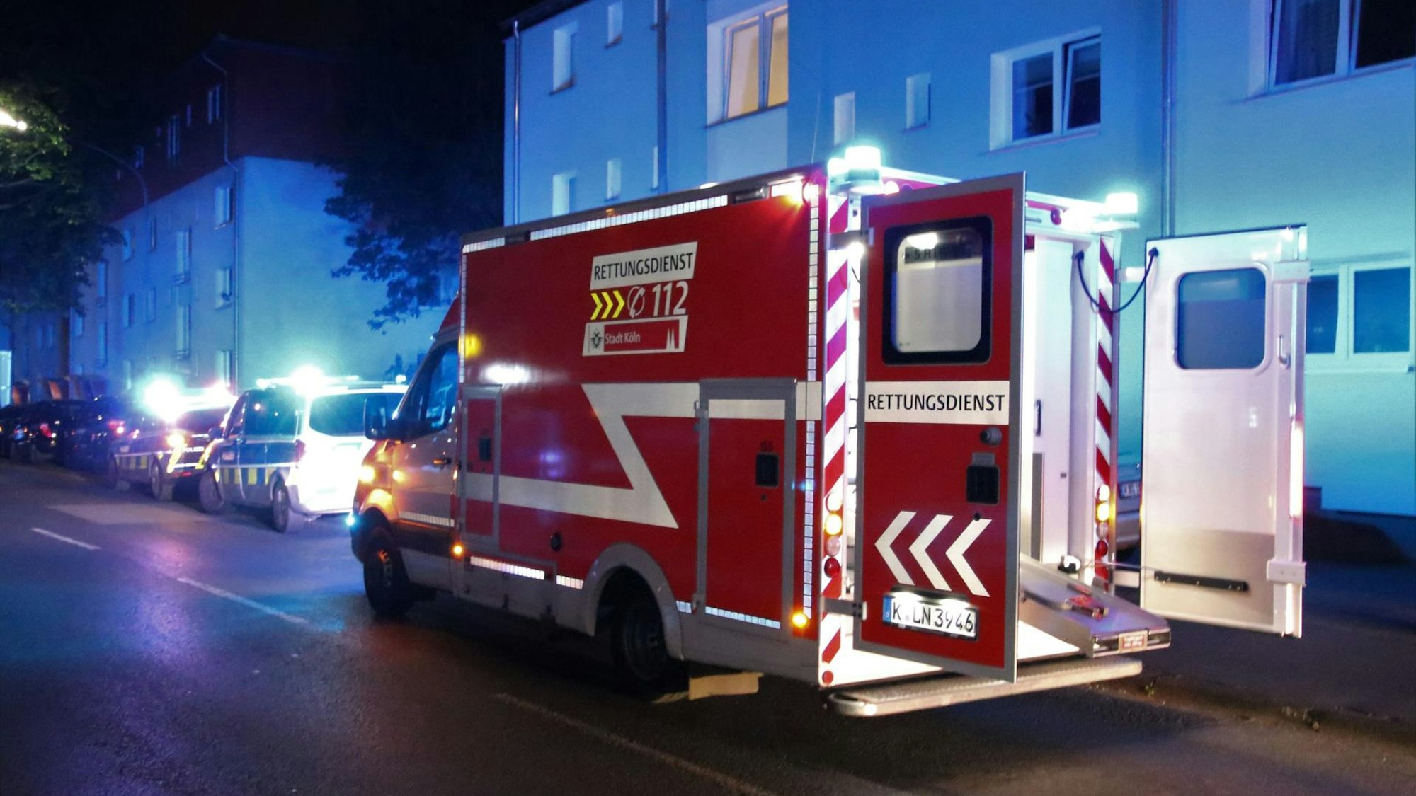 Ein Rettungswagen mit offenen Türen sowie zwei Streifenwagen stehen vor einem Mehrfamilienhaus.