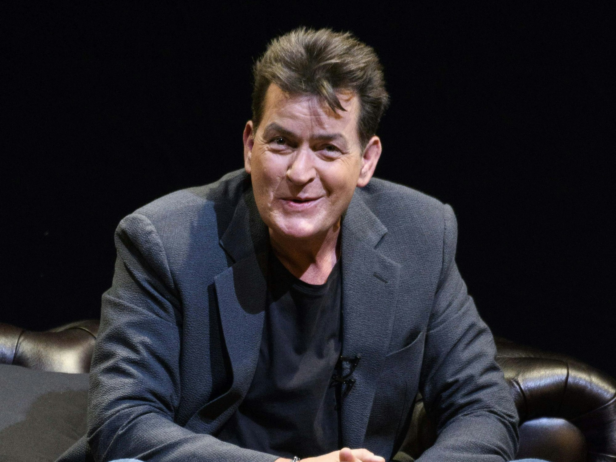 Charlie Sheen sitzt während einer Pressekonferenz gelassen auf einem Ledersofa.