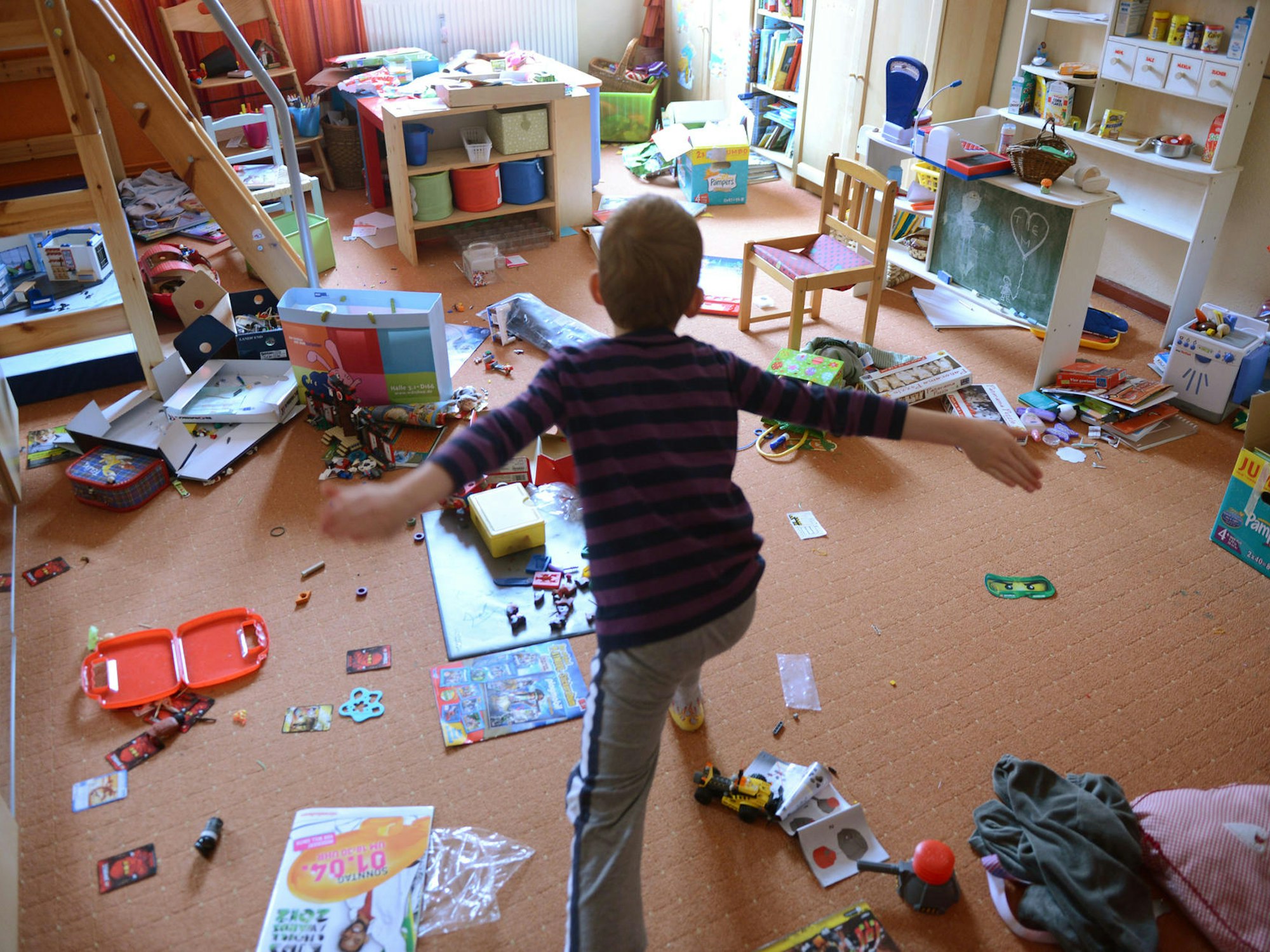 ein Kind tobt in einem Zimmer, in dem zahlreiche Spielsachen wild auf dem Boden liegen.