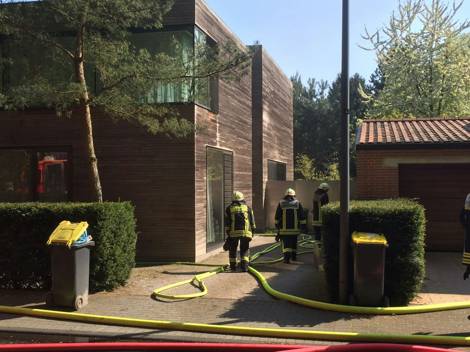 Drei Einsatzkräfte der Feuerwehr stehen vor einem mit Holz verkleidetem Haus, Wasserschläuche liegen in der Einfahrt.