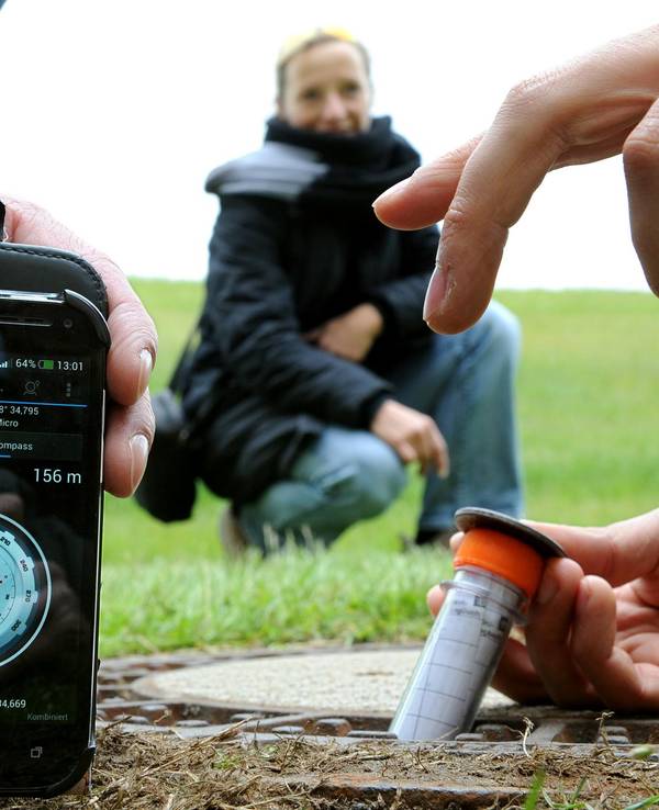 Ein Mann zeigt auf sein Handy, ein zweiter zieht ein Röhrchen aus dem Loch eines Gullideckels, ein Frau guckt zu.