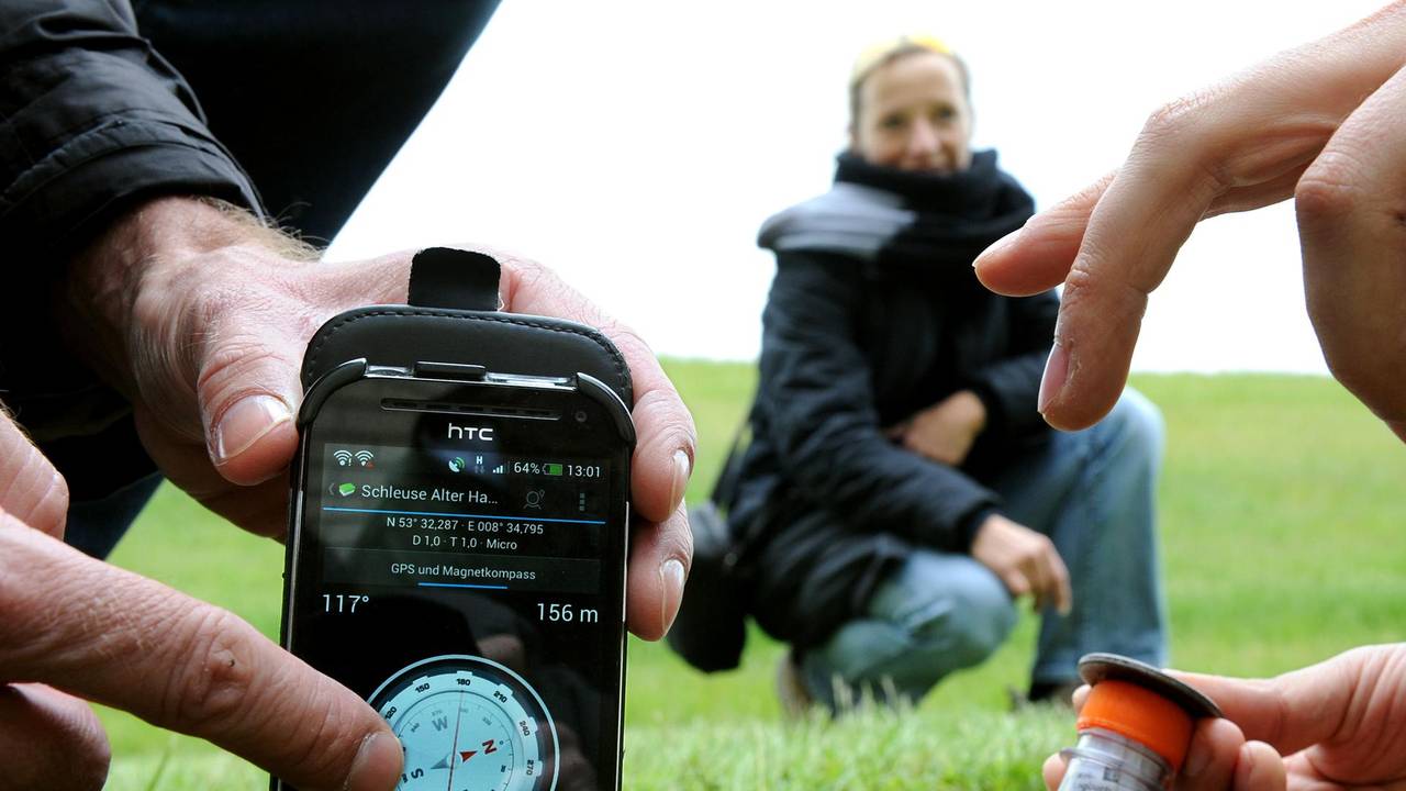 Ein Mann zeigt auf sein Handy, ein zweiter zieht ein Röhrchen aus dem Loch eines Gullideckels, ein Frau guckt zu.