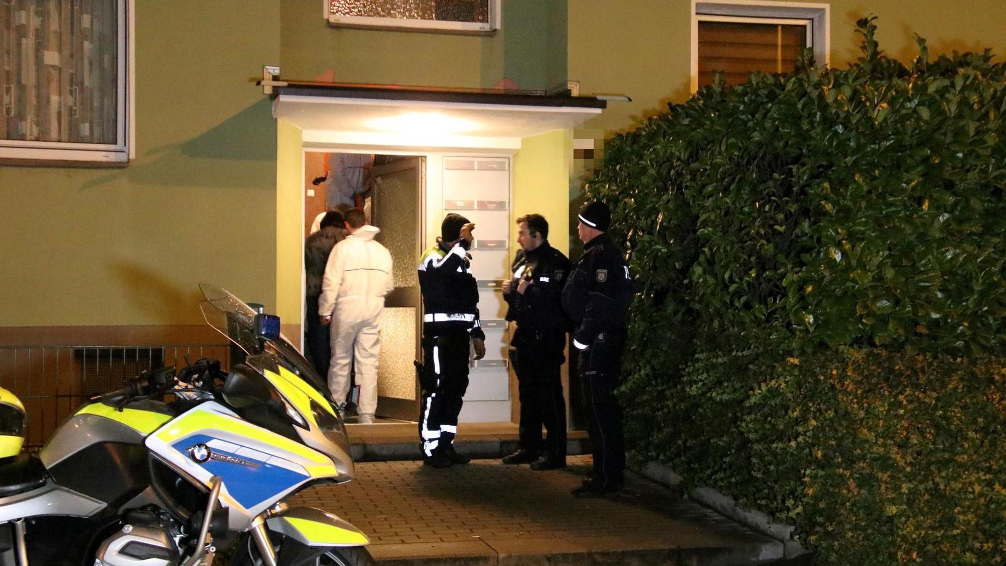 Drei Polizisten stehen vor einem Mehrfamilienhaus, ein Beamter der Spurensicherung in einem Schutzanzug geht in das Haus.