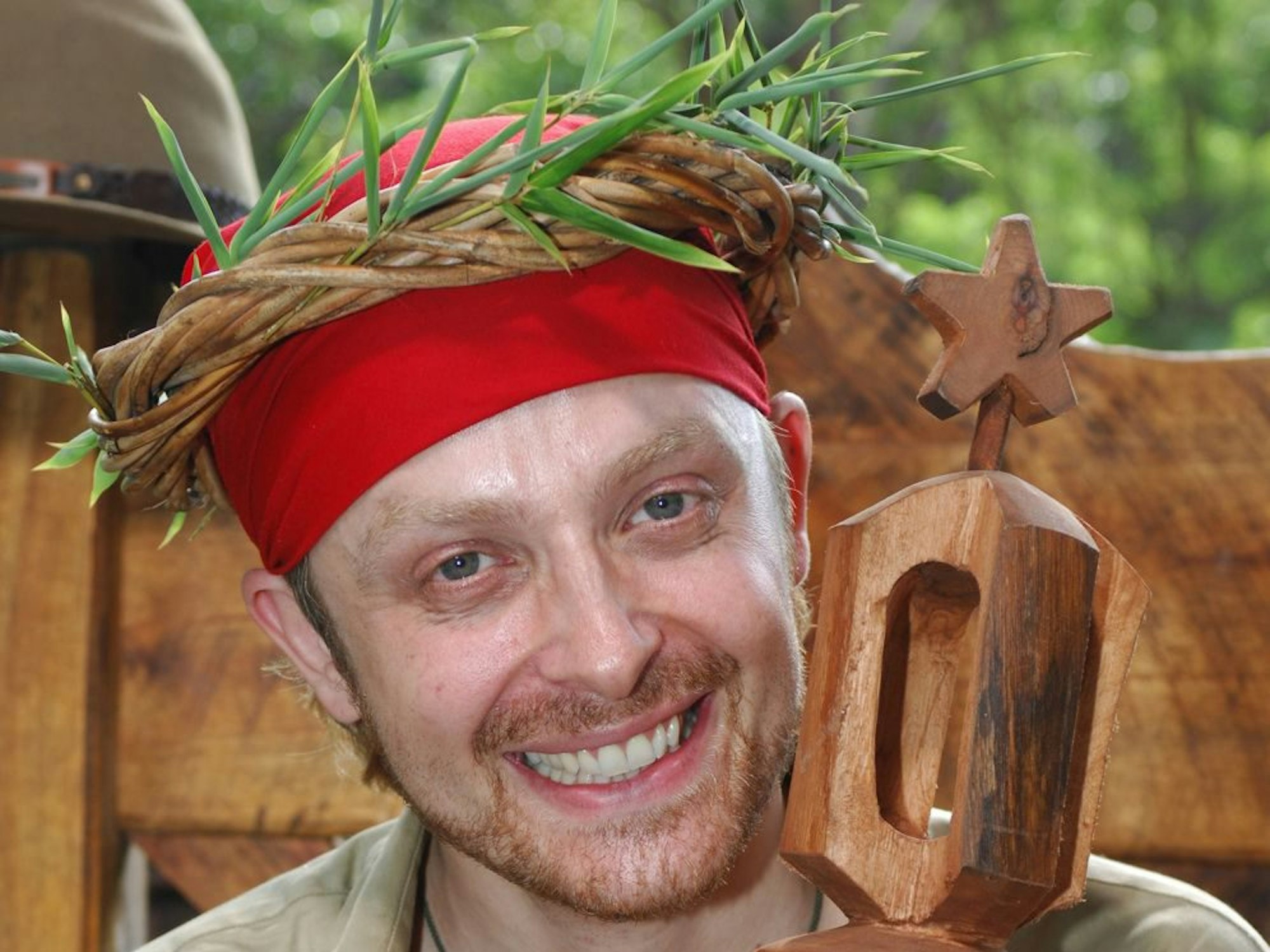 Ein junger Mann mit müdem Gesichtsausdruck trägt eine aus Ästen geflochtene Krone auf dem Kopf und hält ein Zepter in der Hand.