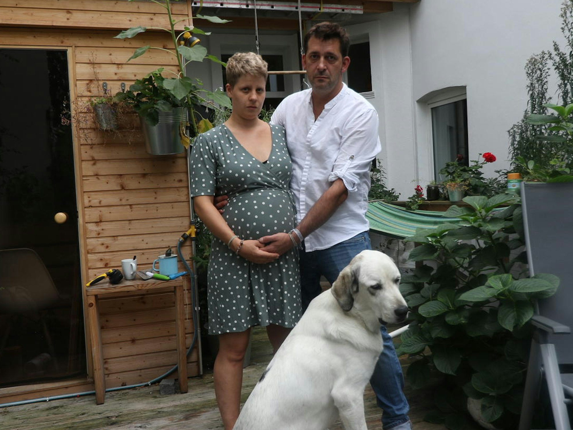 Eine schwangere Frau steht mit einem Mann und Hund auf einer Terrasse.