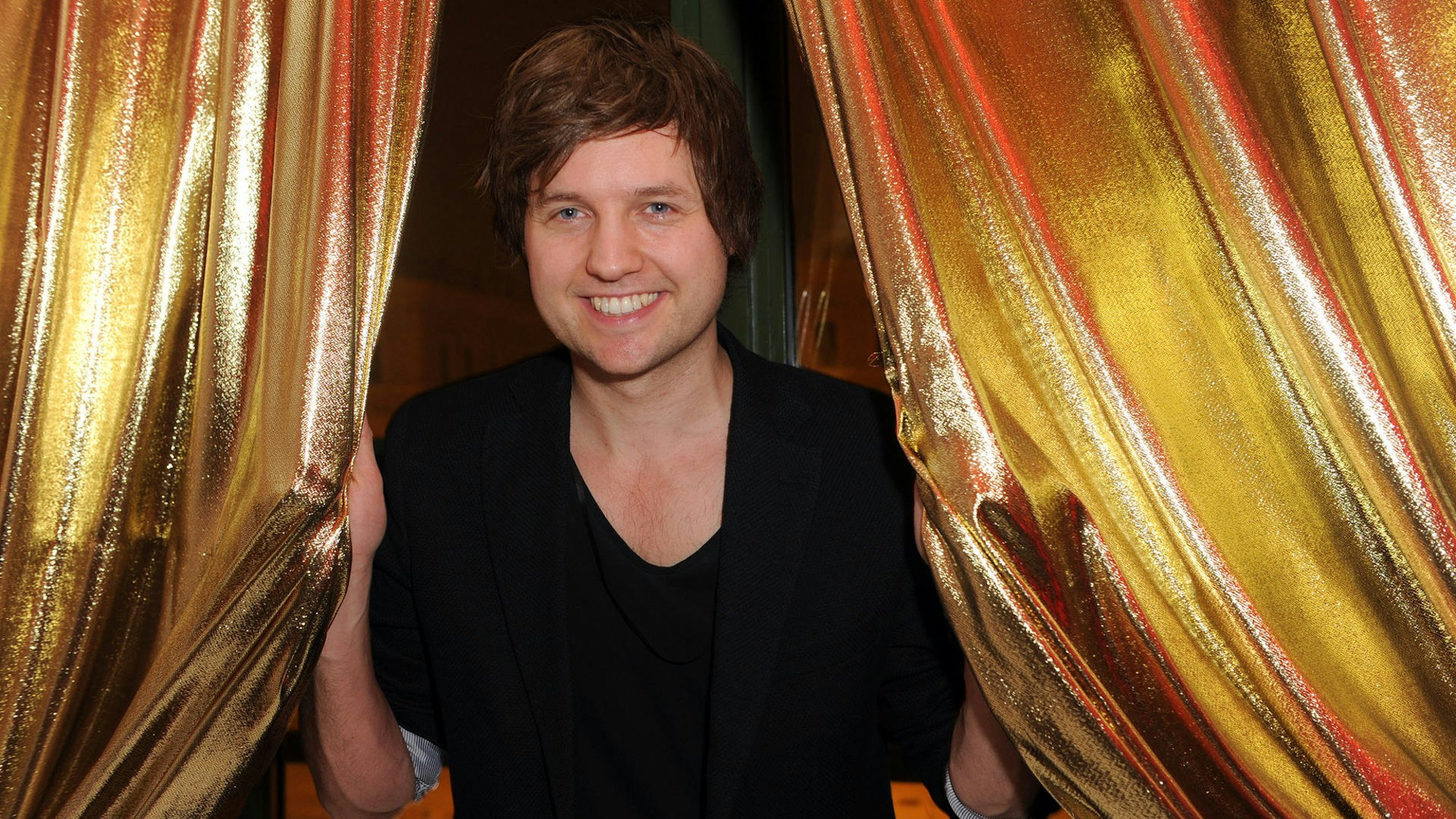 Nick Howard lächelt vor einem goldenen Vorhang in die Kamera.