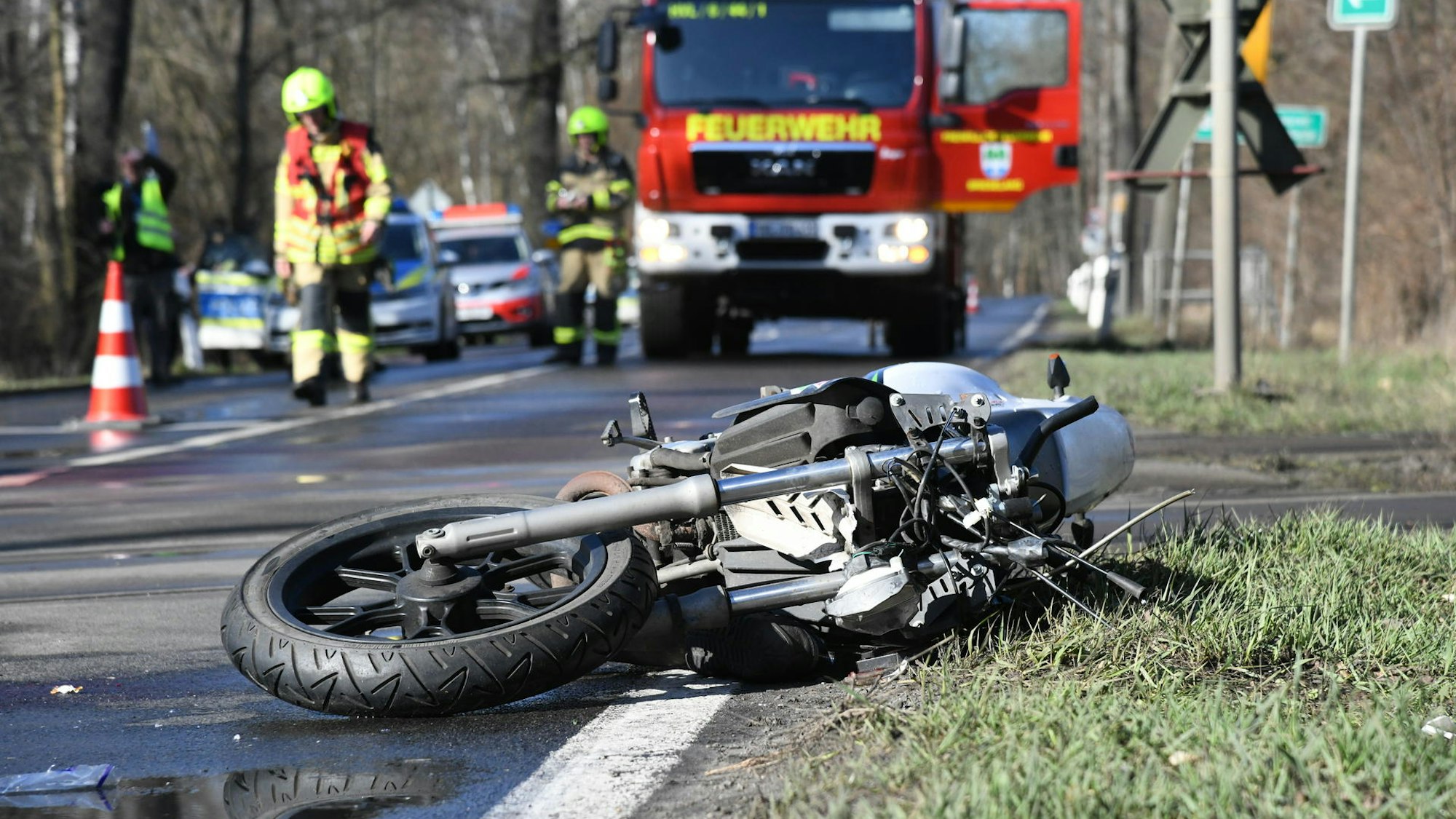 Ein verunglücktes Motorrad liegt auf der Seite an einer Straße, im Hintergrund stehen Einsatzfahrzeuge von Feuerwehr und Polizei sowie Einsatzkräfte.