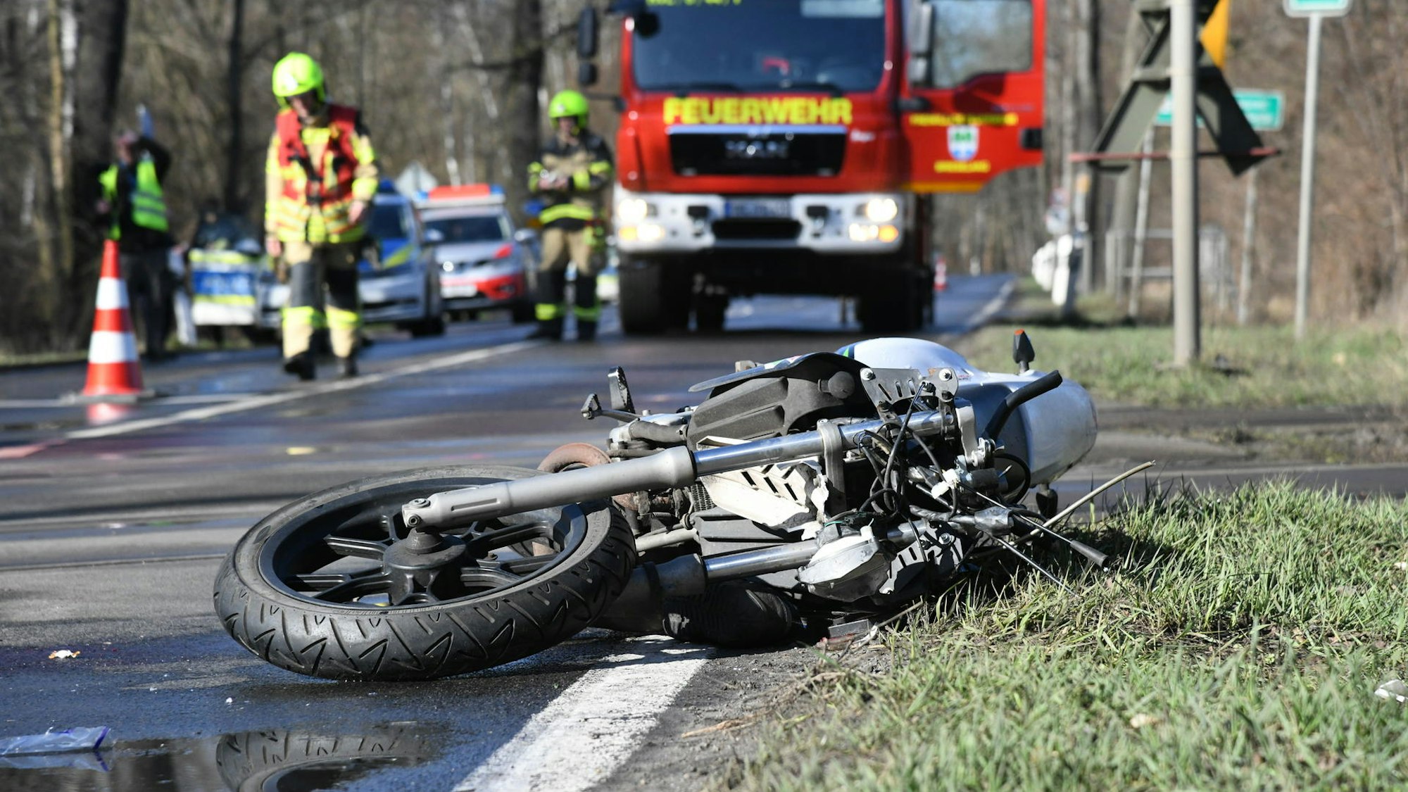 Ein beschädigtes Motorrad liegt halb auf der Fahrbahn, halb im Grünstreifen, im Hintergrund stehen Einsatzfahrzeuge von Feuerwehr und Polizei. Zwei Feuerwehrmänner laufen über die Straße.
