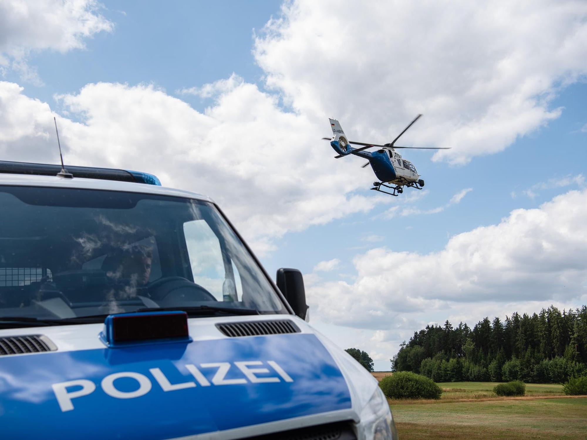 Ein Polizeifahrzeug steht auf einer ländlichen Straße, im Hintergrund fliegt ein Hubschrauber.