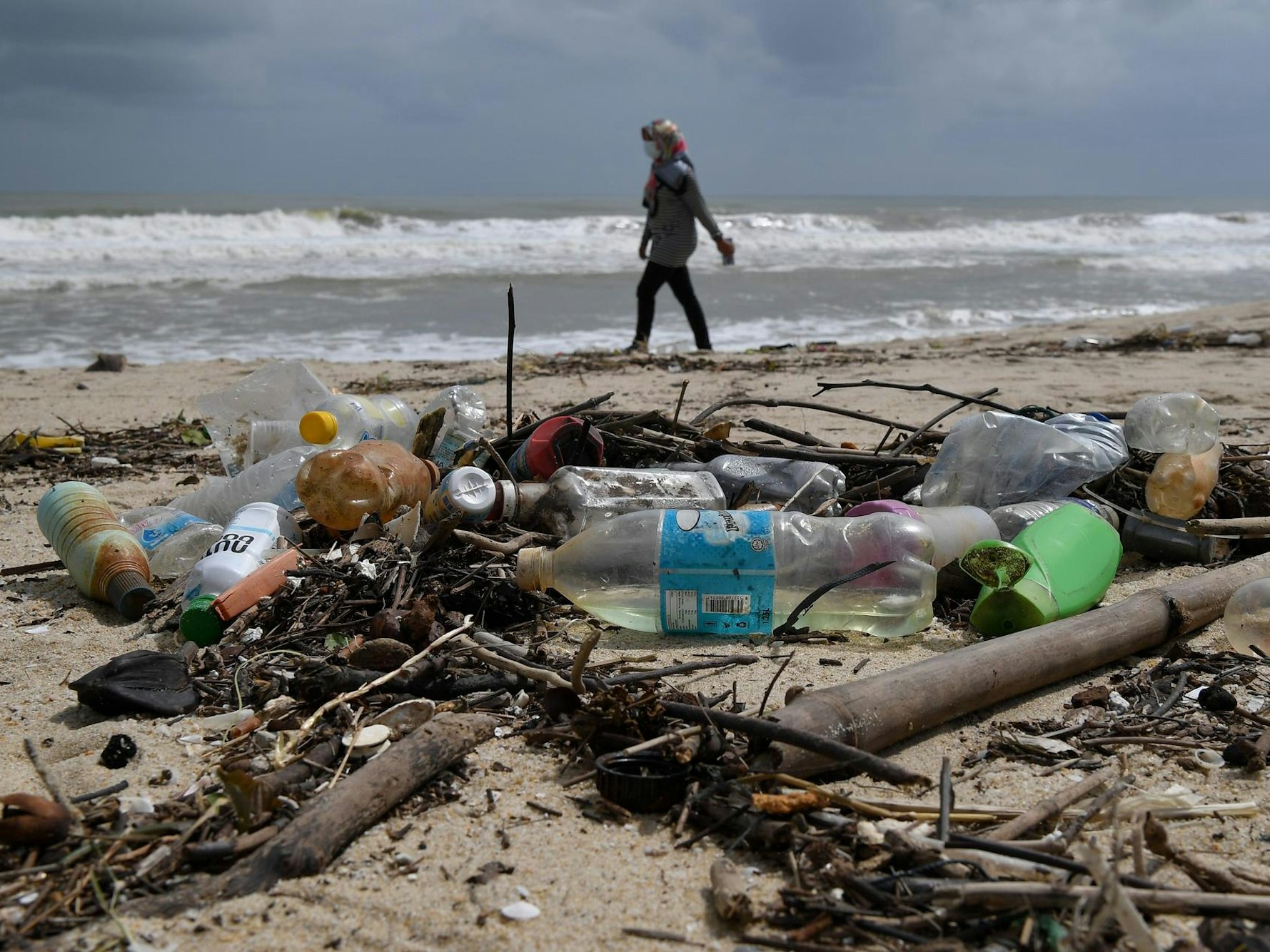Frau geht am Strand spazieren, im Vordergrund liegen Plastikflaschen.