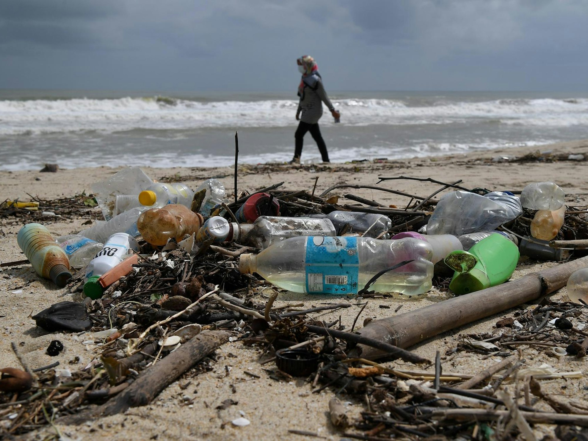 Frau geht am Strand spazieren, im Vordergrund liegen Plastikflaschen.