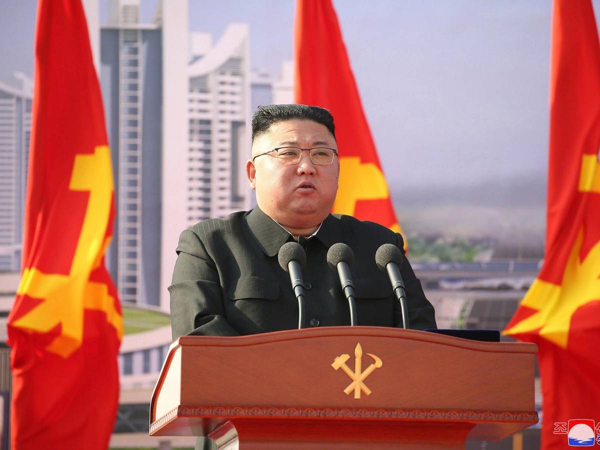 Auf dem Symbolfoto von 2021 steht Kim Jong Un hinter einer Holzkanzel mit drei Mikrofonen, im Hintergrund befinden sich drei Flaggen und Hochhäuser.