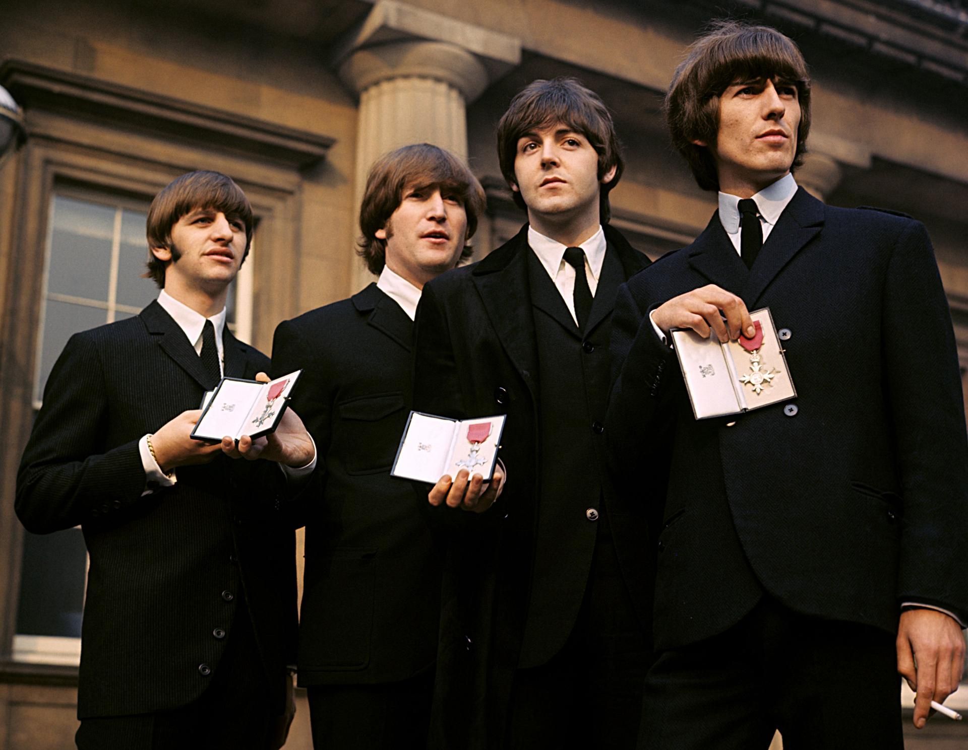 Das undatierte Symbolfoto zeigt die vier Mitglieder der Band „The Beatles“. Sie tragen schwarze Anzüge und halten Pässe mit einer Auszeichnung.