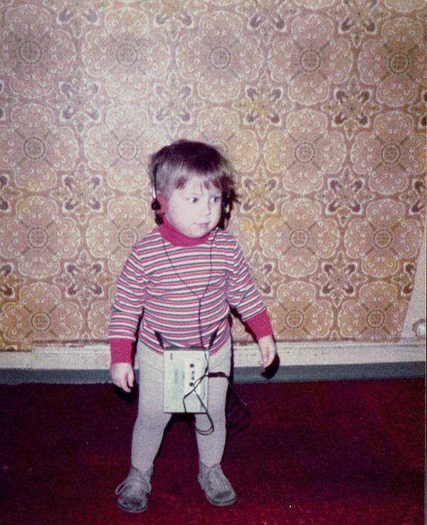 Kerstin Ott als Kind im bunt gestreiften Pullover, mit einem Kopfhörer auf der wuscheligen Frisur, der zugehörige Walkman baumelt an den Beinen.