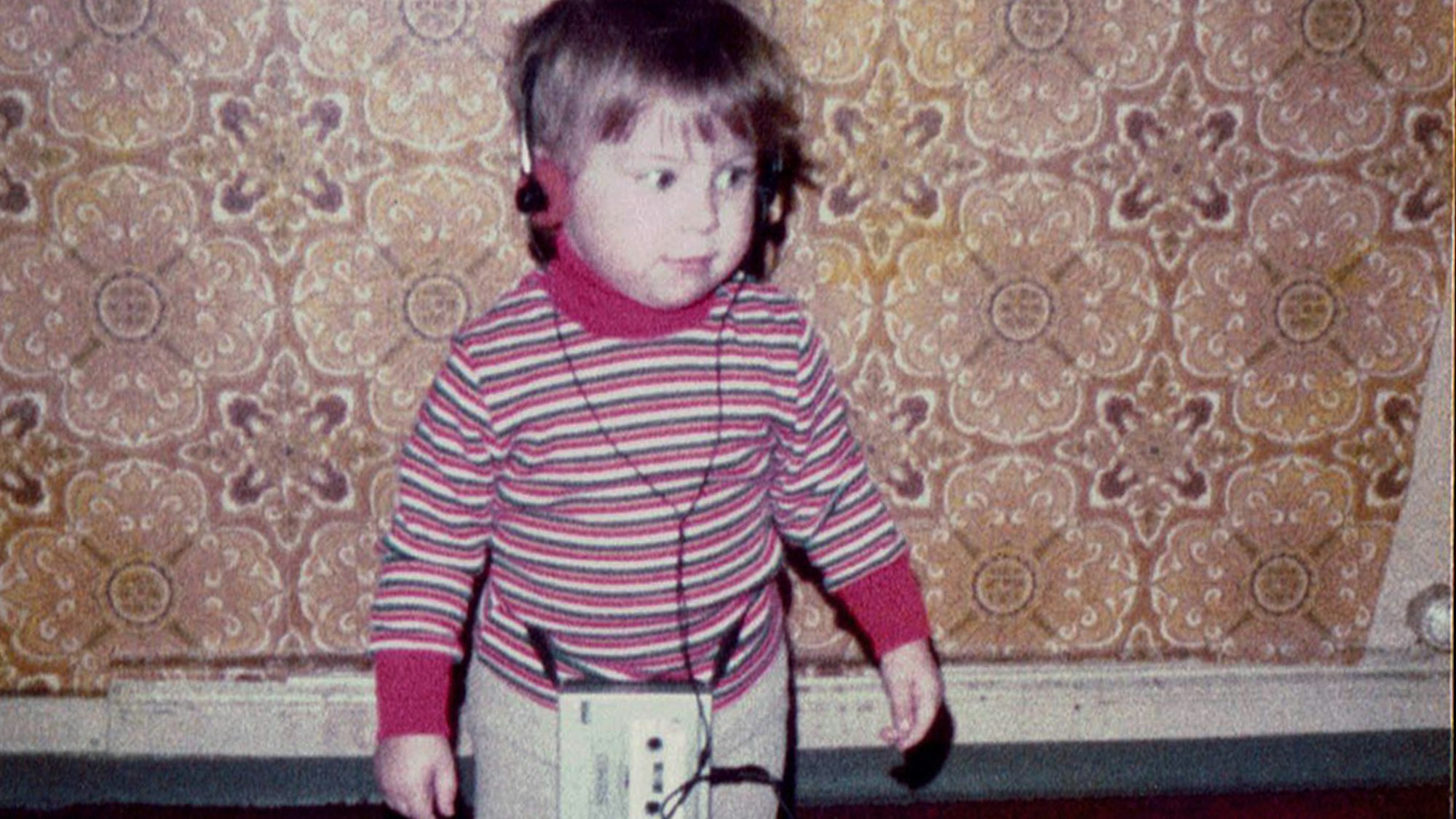 Kerstin Ott als Kind im bunt gestreiften Pullover, mit einem Kopfhörer auf der wuscheligen Frisur, der zugehörige Walkman baumelt an den Beinen.