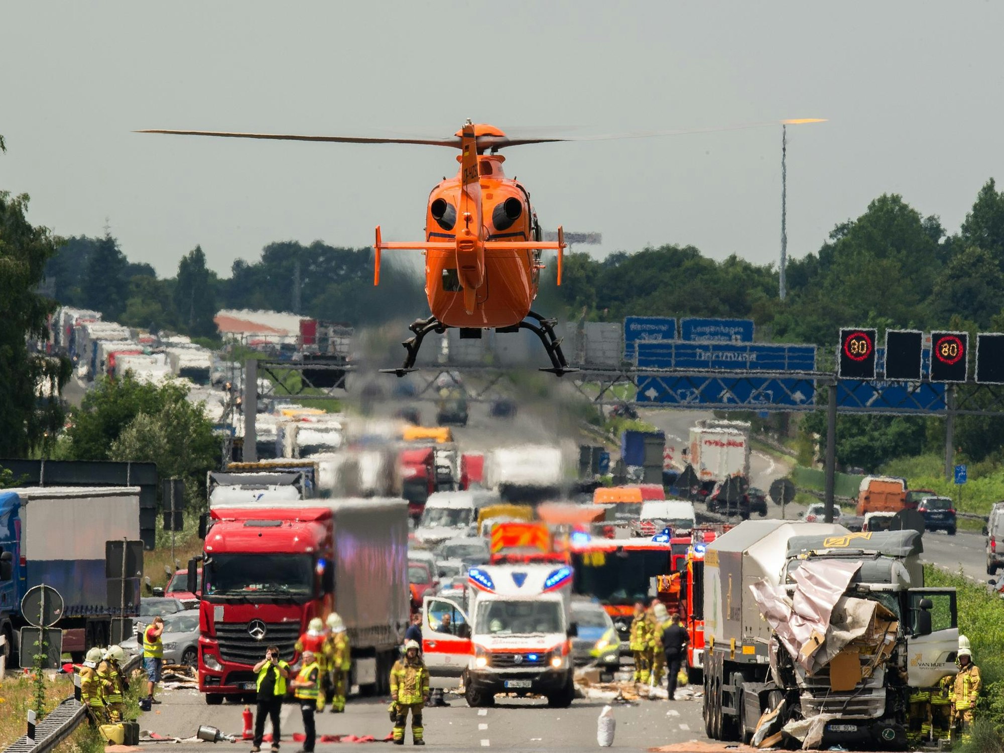 Ein Rettungshubschrauber fliegt zu einer Unfallstelle auf einer Autobahn. Dort stehen zwei beschädigte Lkw, daneben steht ein Rettungswagen, zahlreiche Einsatzkräfte befinden sich auf der Fahrbahn.