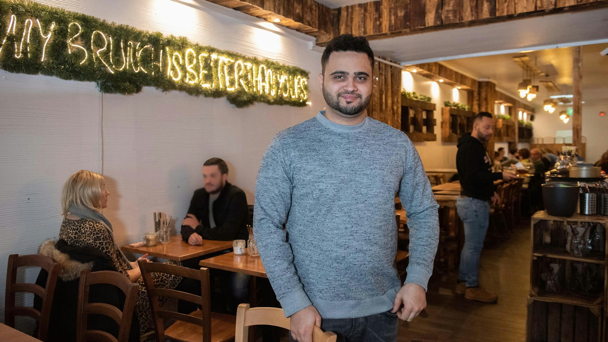 Parham Pooramin steht mit einem grauen Pullover im Café Buur in Köln, im Hintergrund sind Gäste zu sehen.