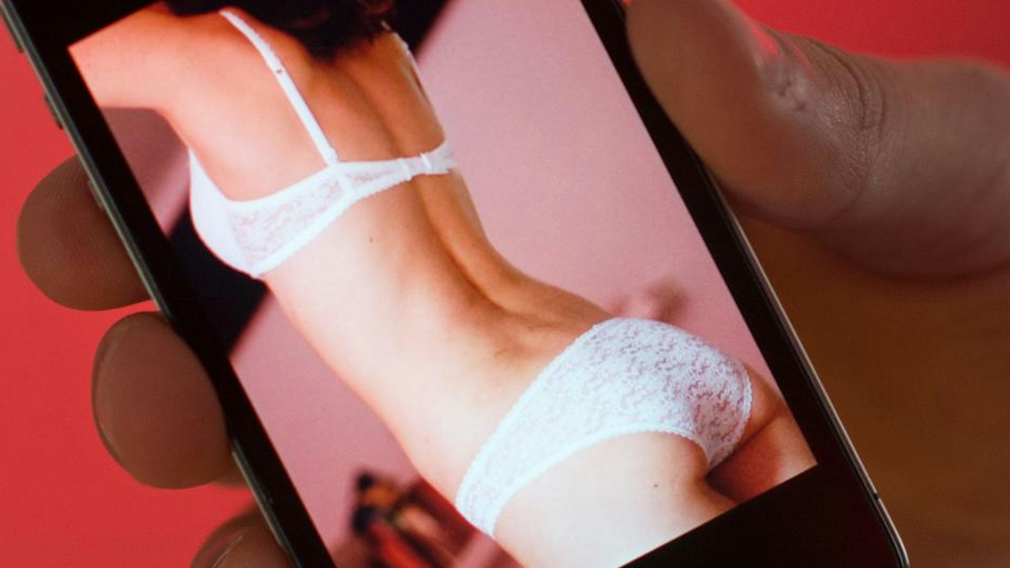 Ein junger Mann hält ein Smartphone, auf dem ein erotisches Foto einer jungen erwachsenen Frau zu sehen ist.