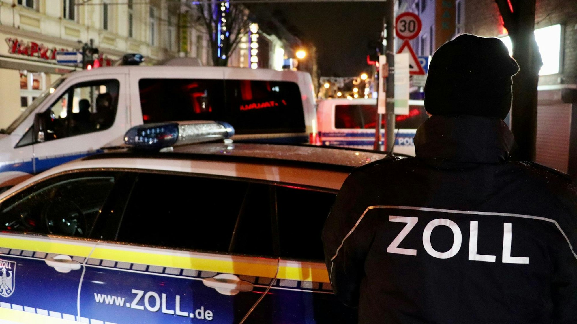 Ein Zollbeamter sowie zwei Zollfahrzeuge stehen an einer Straße.