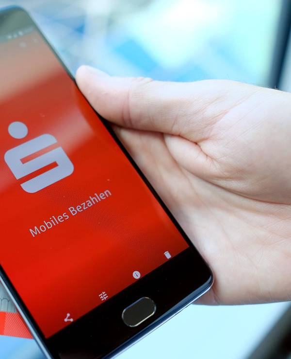 Das undatierte Symbolfoto zeigt eine Hand, die ein schwarzes Smartphone mit der Sparkassen-App hält. Hinter dem Smartphone befindet sich eine EC-Karte der Bank.