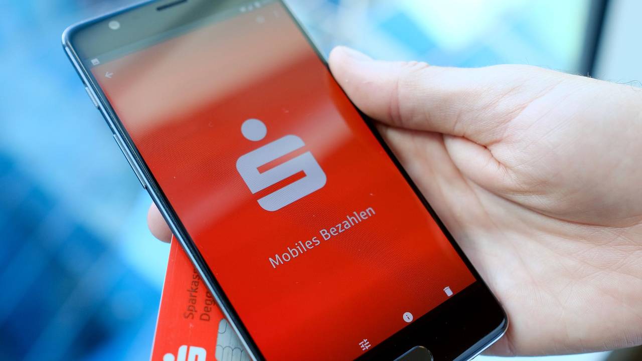 Das undatierte Symbolfoto zeigt eine Hand, die ein schwarzes Smartphone mit der Sparkassen-App hält. Hinter dem Smartphone befindet sich eine EC-Karte der Bank.