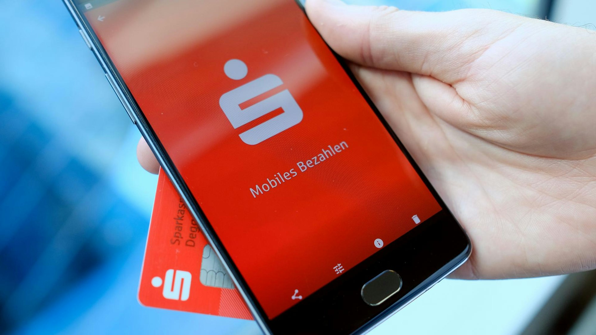 Das undatierte Symbolfoto zeigt eine Hand, die ein schwarzes Smartphone mit der Sparkassen-App hält. Hinter dem Smartphone befindet sich eine EC-Karte der Bank.