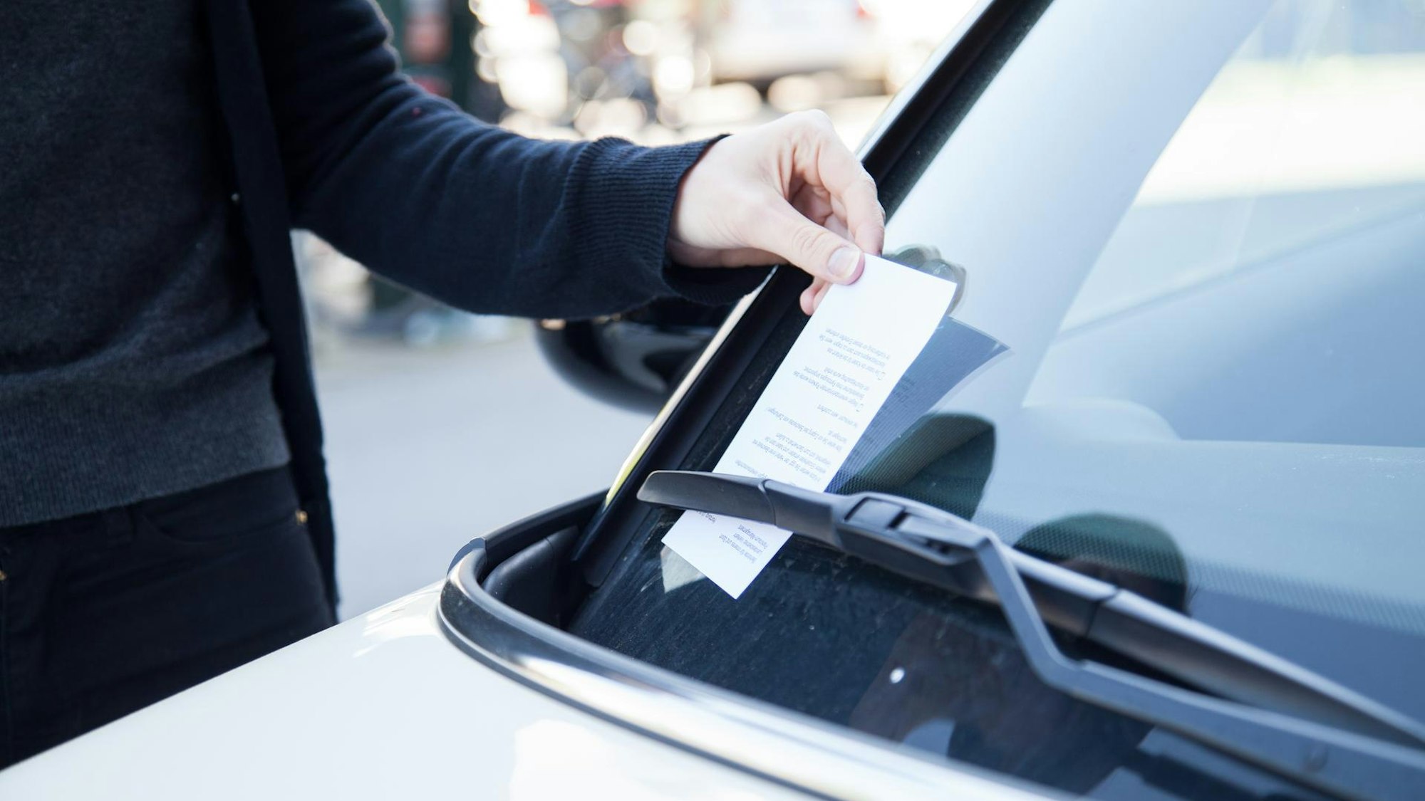Eine Person greift ein Knöllchen, das hinter dem Scheibenwischer eines Fahrzeuges steckt.