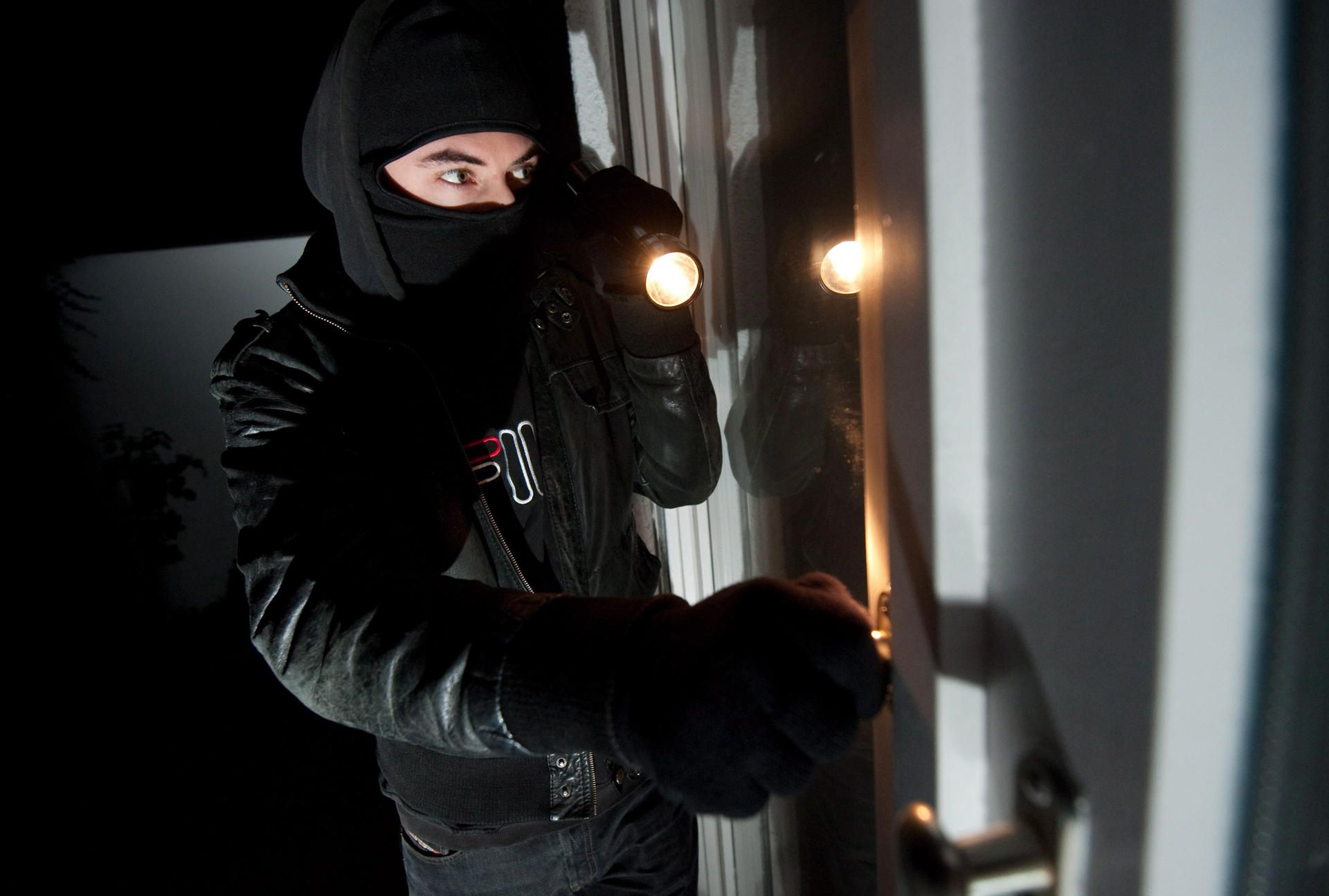 Ein vermummter Einbrecher leuchtet mit einer Taschenlampe in eine Wohnung.