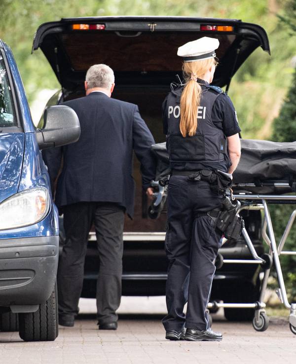 Bestatter bringen einen Leichnam zum Leichenwagen, davor steht eine Polizistin.