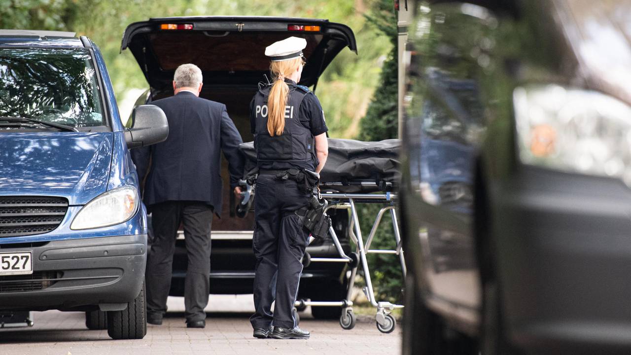 Bestatter bringen einen Leichnam zum Leichenwagen, davor steht eine Polizistin.