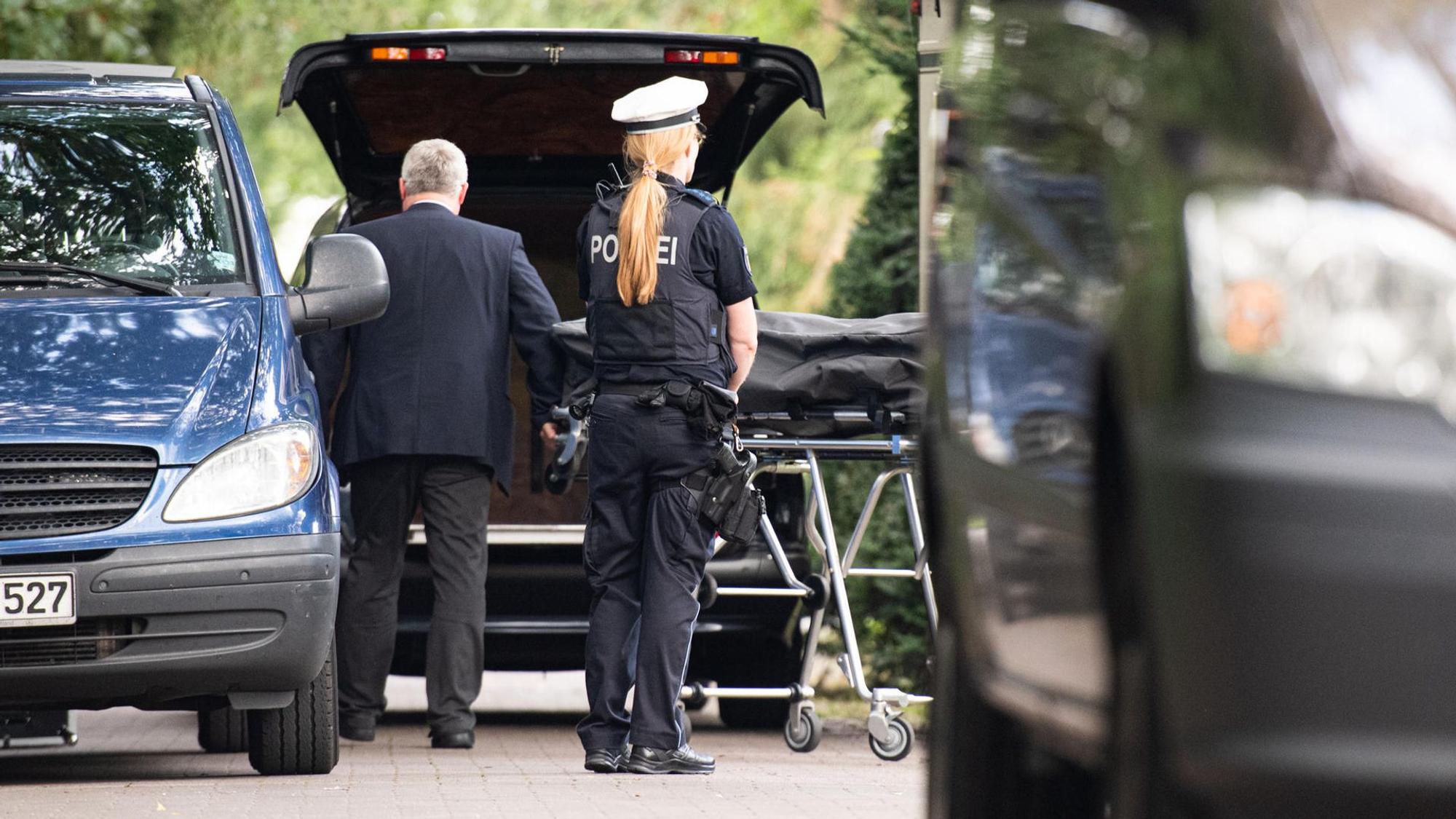 Bestatter bringen einen Leichnam zum Leichenwagen, davor steht eine Polizistin.