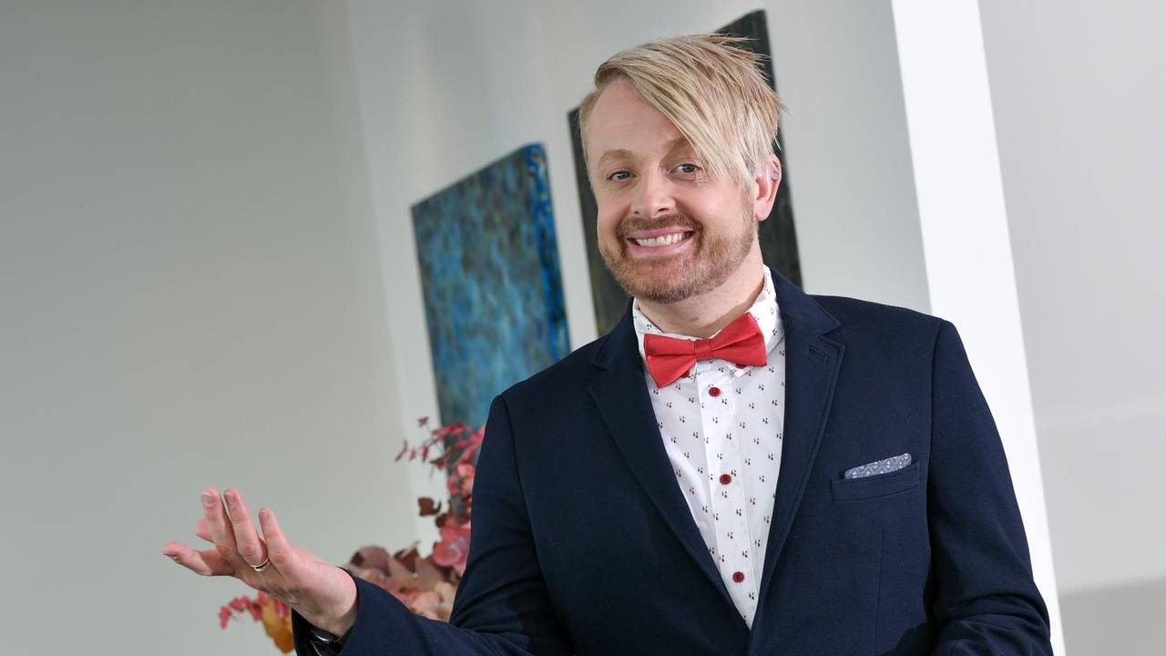 Ein blonder junger Mann mit Jackett und Fliege macht eine einladende Handbewegung.