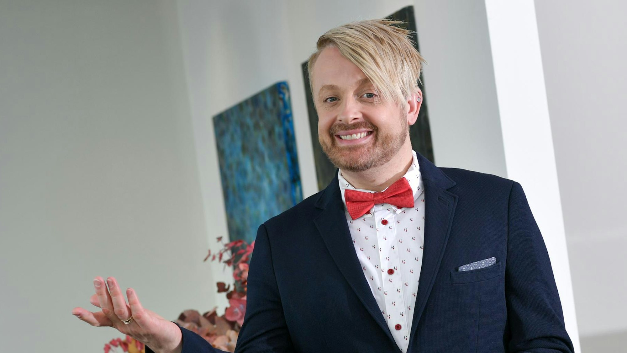 Ein blonder junger Mann mit Jackett und Fliege macht eine einladende Handbewegung.