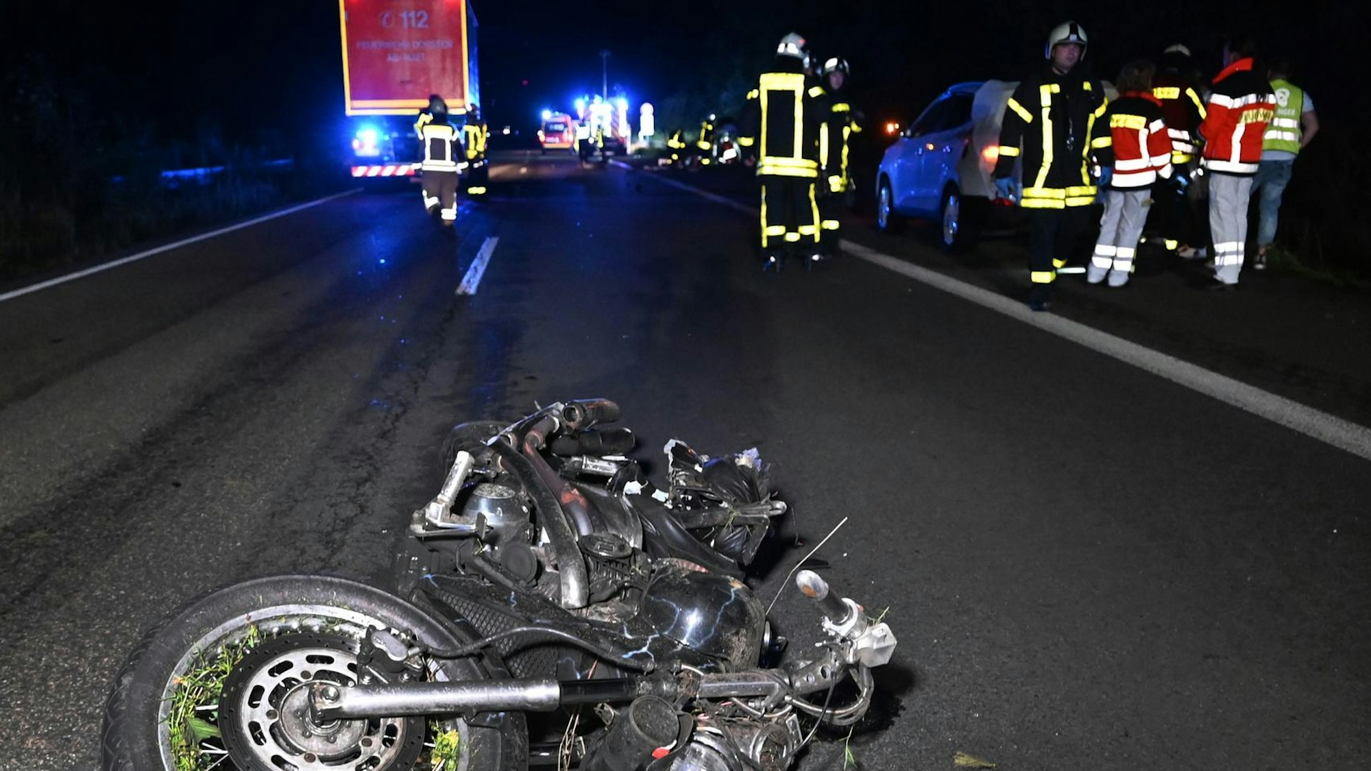 Ein zerfetztes Motorrad liegt auf der Fahrbahn, im Hintergrund stehen beziehungsweise laufen Einsatzkräfte von Feuerwehr und Rettungsdienst.