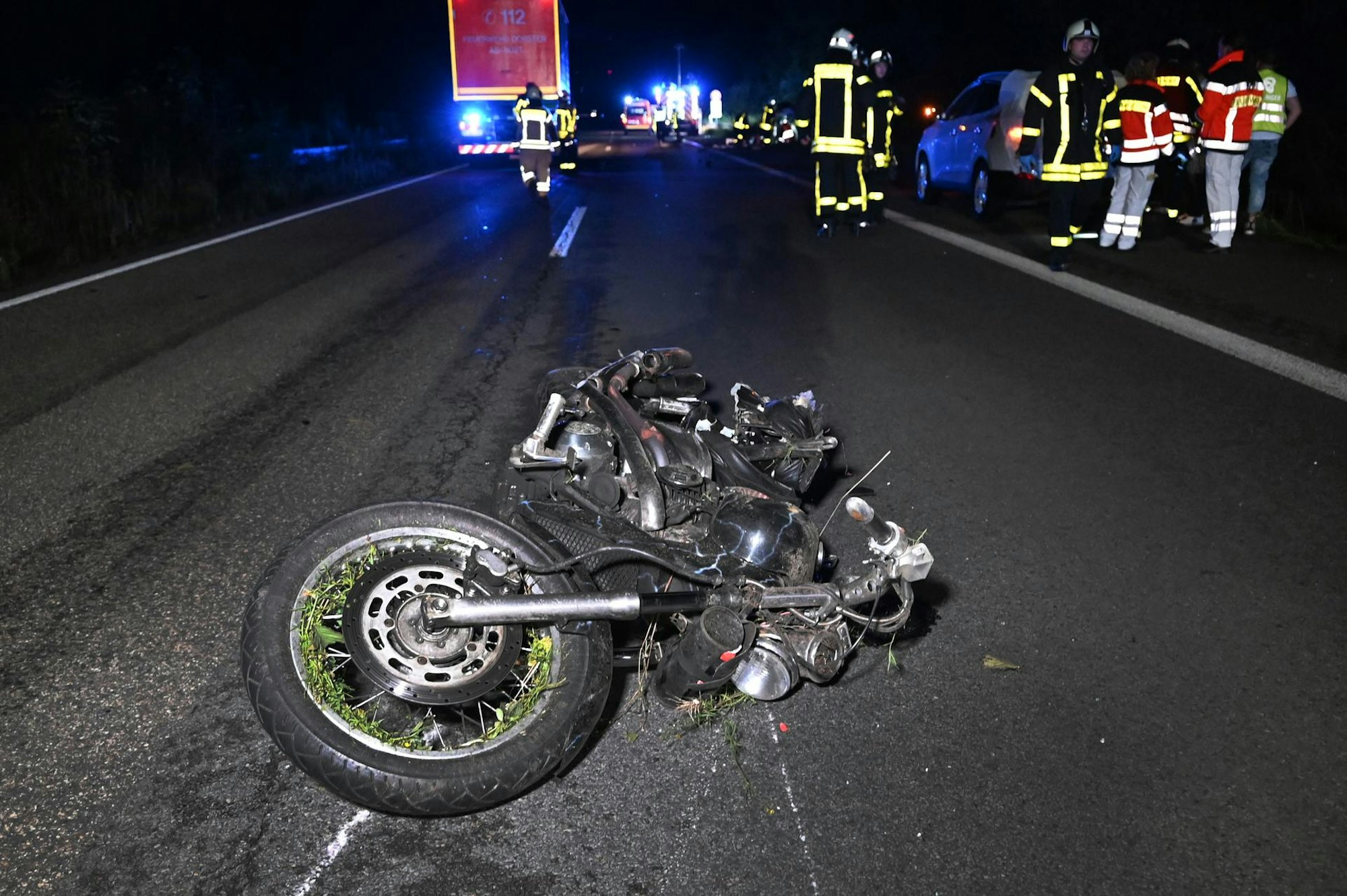 Ein beschädigtes Motorrad liegt auf der Fahrbahn, dahinter zahlreiche Einsatzkräfte von Feuerwehr, Rettungsdienst und Polizei.