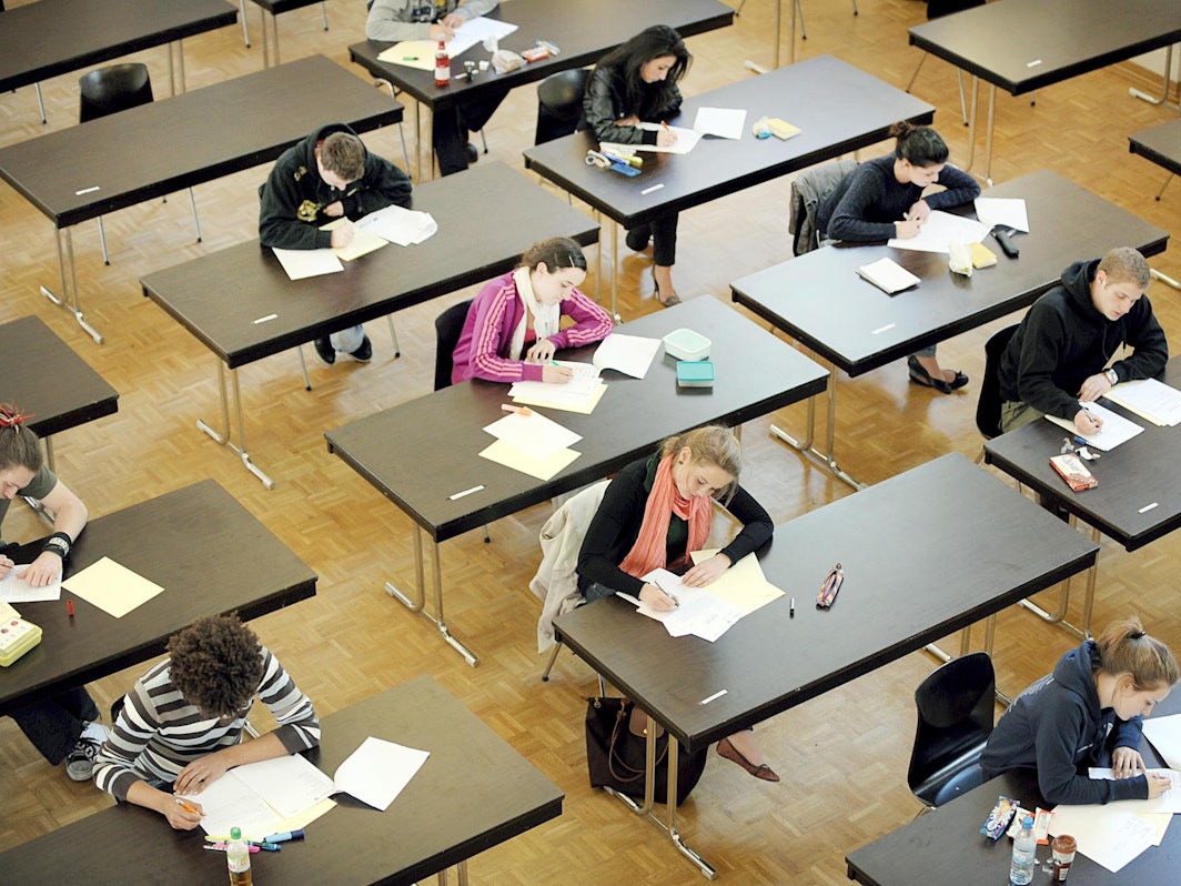 Junge Frauen und Männer sitzen bei einer Prüfung weit auseinander an Tischen.