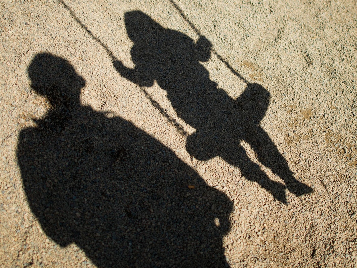 Der Schatten von einem Mann und einem schaukelnden Kind fallen auf Sand auf einem Spielplatz.