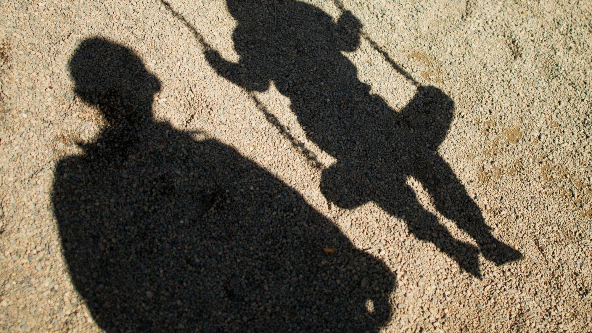 Der Schatten von einem Mann und einem schaukelnden Kind fallen auf Sand auf einem Spielplatz.
