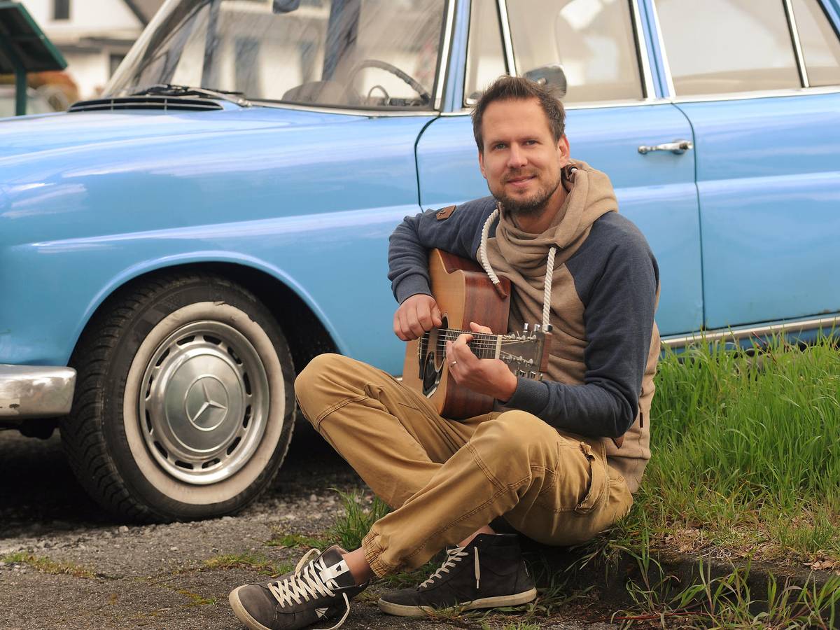 Mike Kremer von Miljö spielt sitzend Gitarre vor einem Auto